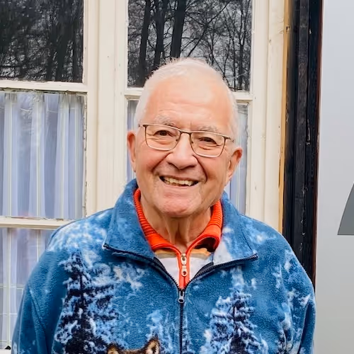 Älterer Mann mit Brille lächelt vor einem Fenster, trägt blauen Pullover mit Waldmotiv.