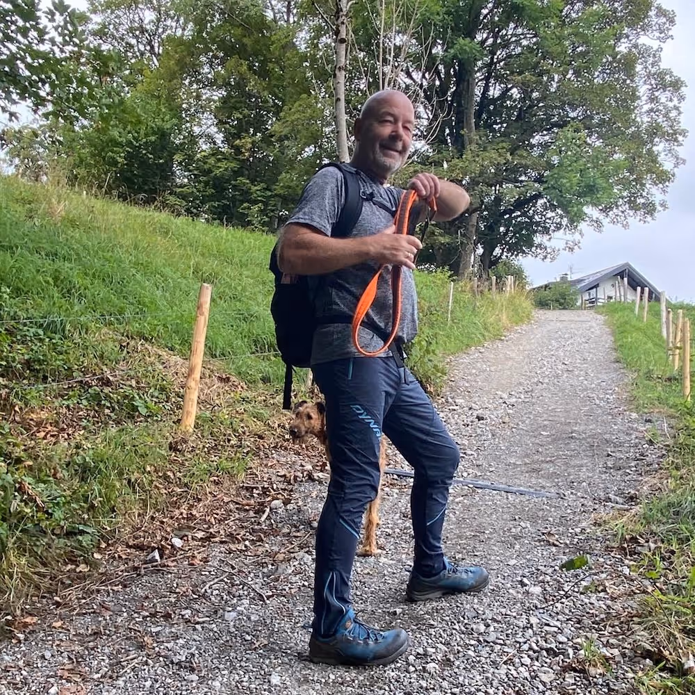 Mann mit Rucksack hält orangefarbene Hundeleine auf einem Wanderweg neben einem braunen Hund.
