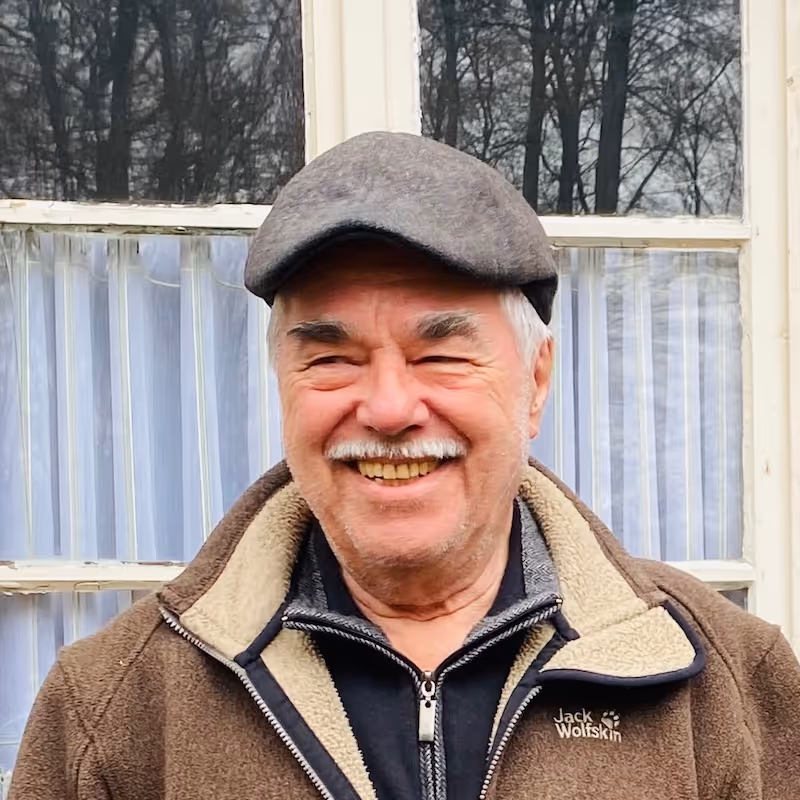 Älterer Mann mit grauer Schirmmütze und brauner Jacke lächelt vor einem Fenster.