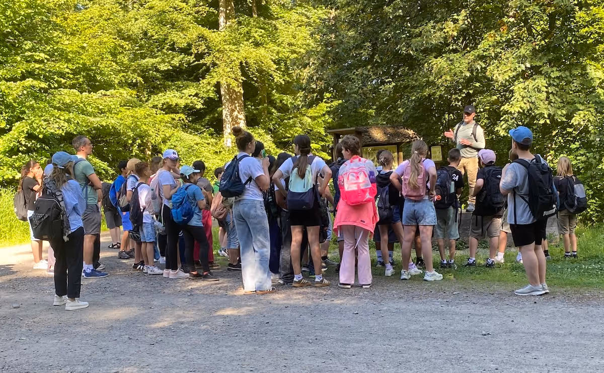 Gruppe von Kindern mit Rucksäcken hört bei sonnigem Waldspaziergang einem Mann mit Kappe zu.