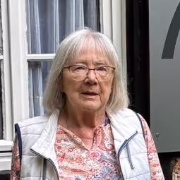 Ältere Frau mit grauem Haar, Brille und weißer Weste steht vor einem Hausfenster.