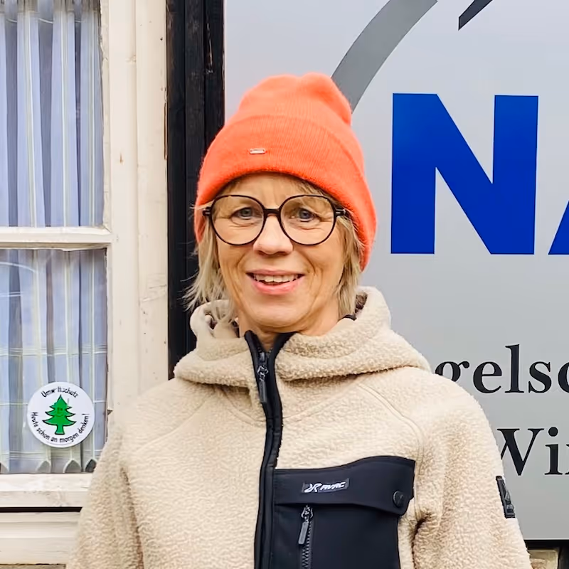 Frau mit orangefarbener Mütze und Brille, trägt eine beige Fleecejacke und lächelt.